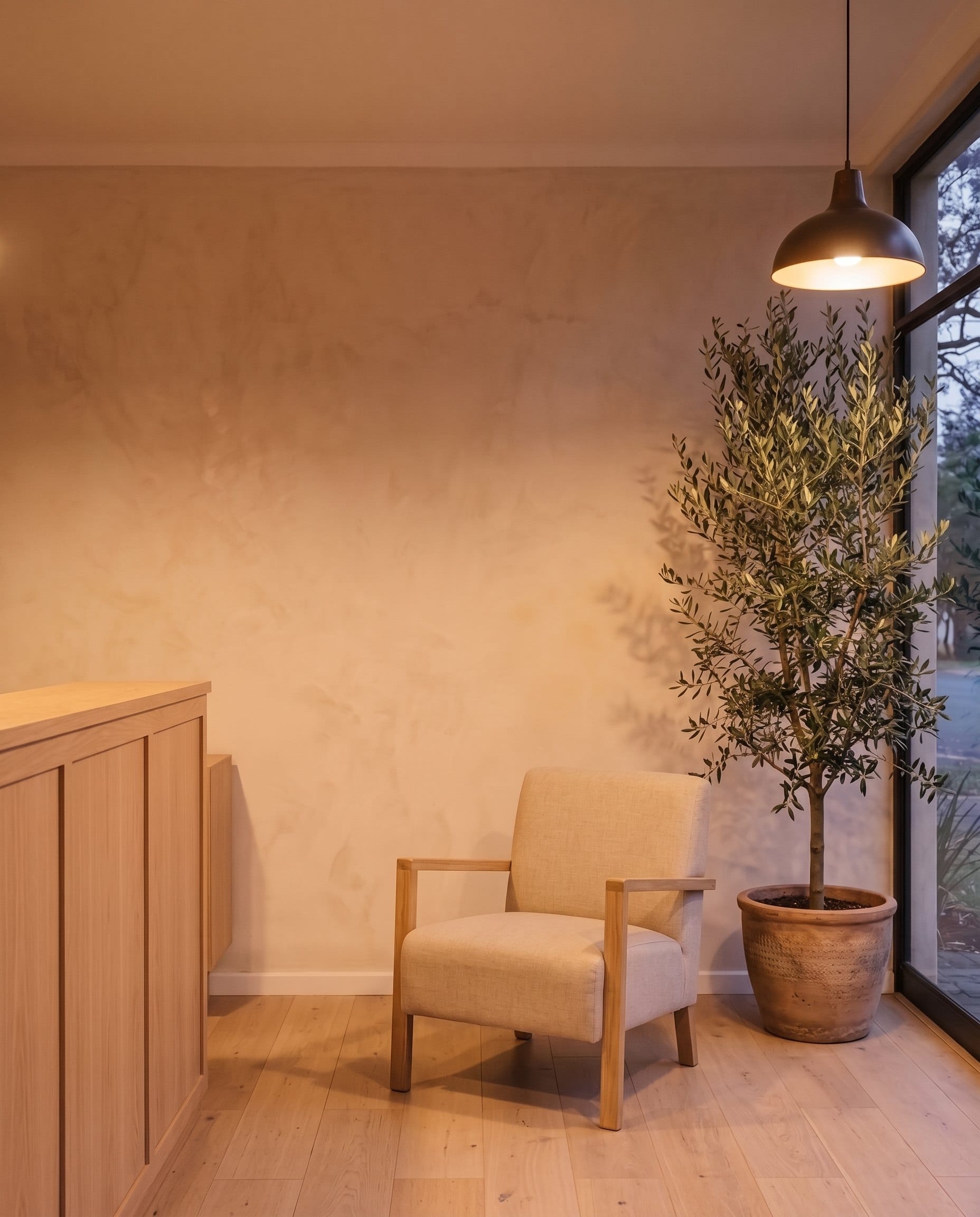 Inside a calm, empty waiting room at dusk: an armchair, an olive tree and warm pendant light
